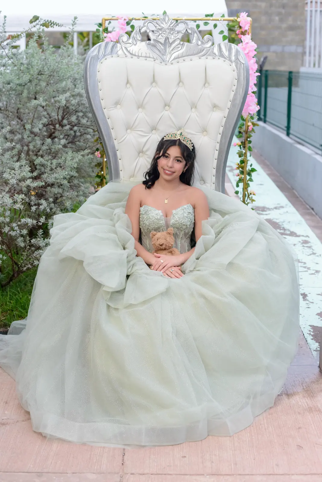 Quinceañera sentada en trono blanco con vestido verde salvia y arco floral rosado